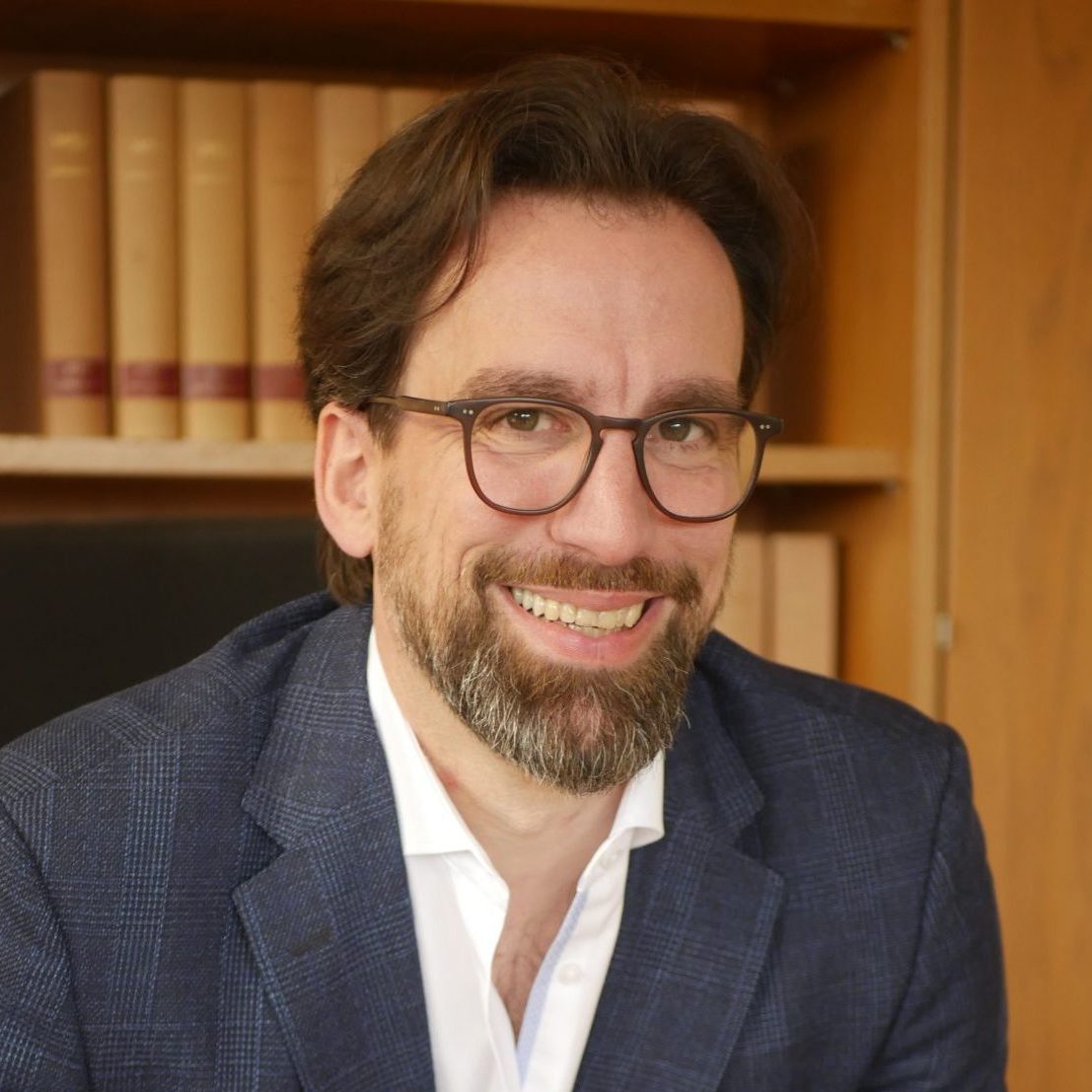 Lächelnder Anwalt Torsten Kowalewski mit Brille, Bart und einer Anzug-Jacke vor einem Bücherregal